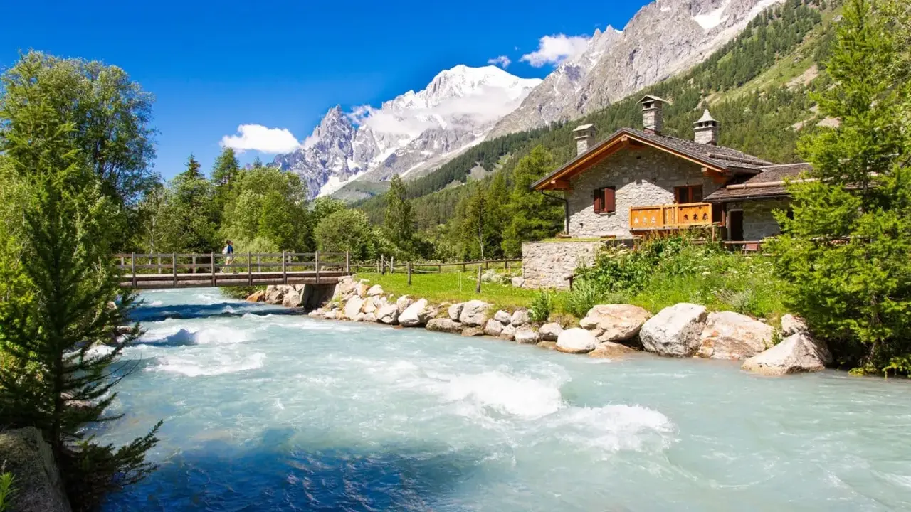 Val Ferret: un'incantevole casa in pietra alpina vicino a un fiume turchese, con un ponte e montagne innevate sullo sfondo.