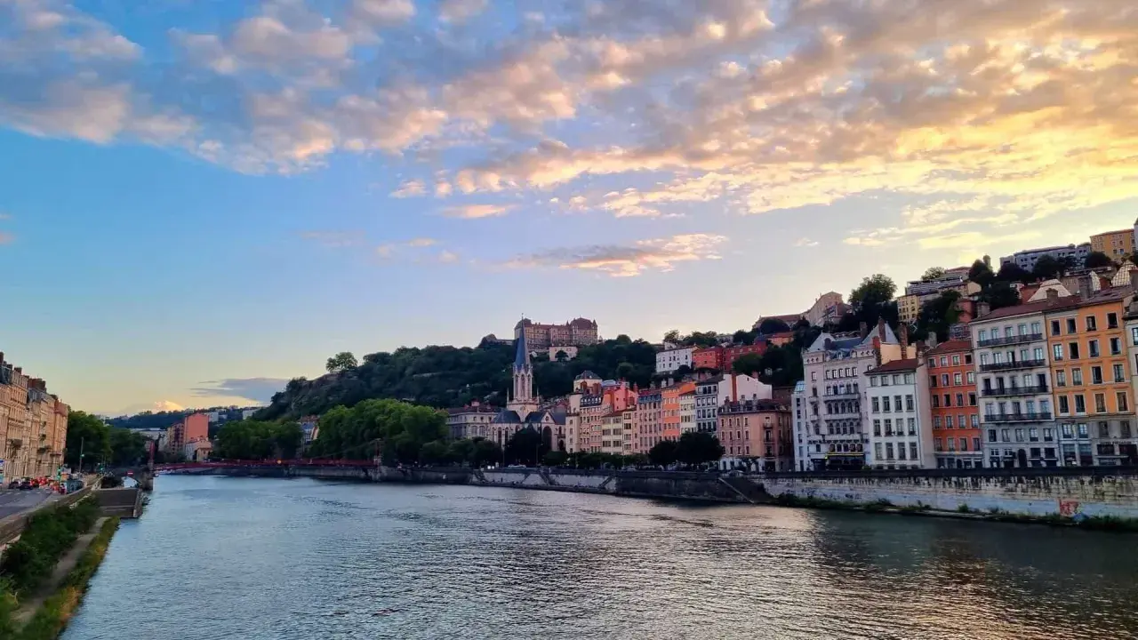 Blick auf die Stadt Lyon, Frankreich, mit dem Fluss Saône im Vordergrund und bunten Gebäuden, die sich den Hügel hinaufziehen. Ein Schloss thront über der Stadt.