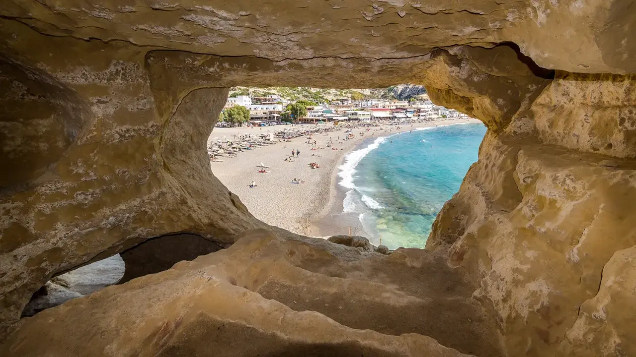 Blick durch Höhle auf Strand von Matala, Kreta. Ein sonniges Hippie-Dorf mit bunten Häusern und blauem Meer.