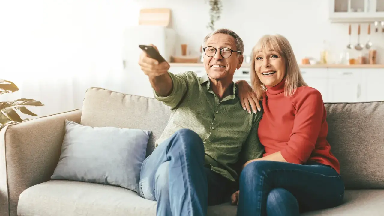 Uśmiechnięta para seniorów ogląda telewizję. Zastanawiają się, od jakiego wieku nie płaci się abonamentu RTV, ciesząc się wspólnym czasem.