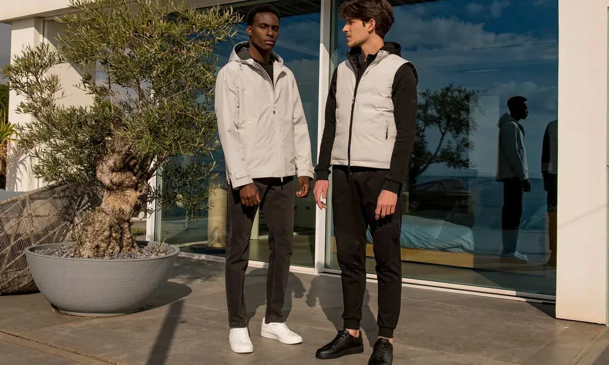 Due ragazzi con look autunnali, uno con giacca leggera, l'altro con gilet imbottito, posano vicino a un ulivo.