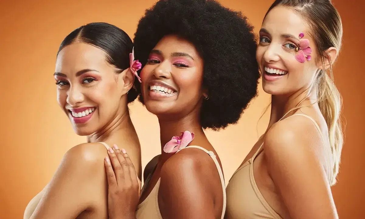 Trois femmes souriantes, un vrai "glow up traduction" avec des fleurs dans les cheveux et un maquillage éclatant.