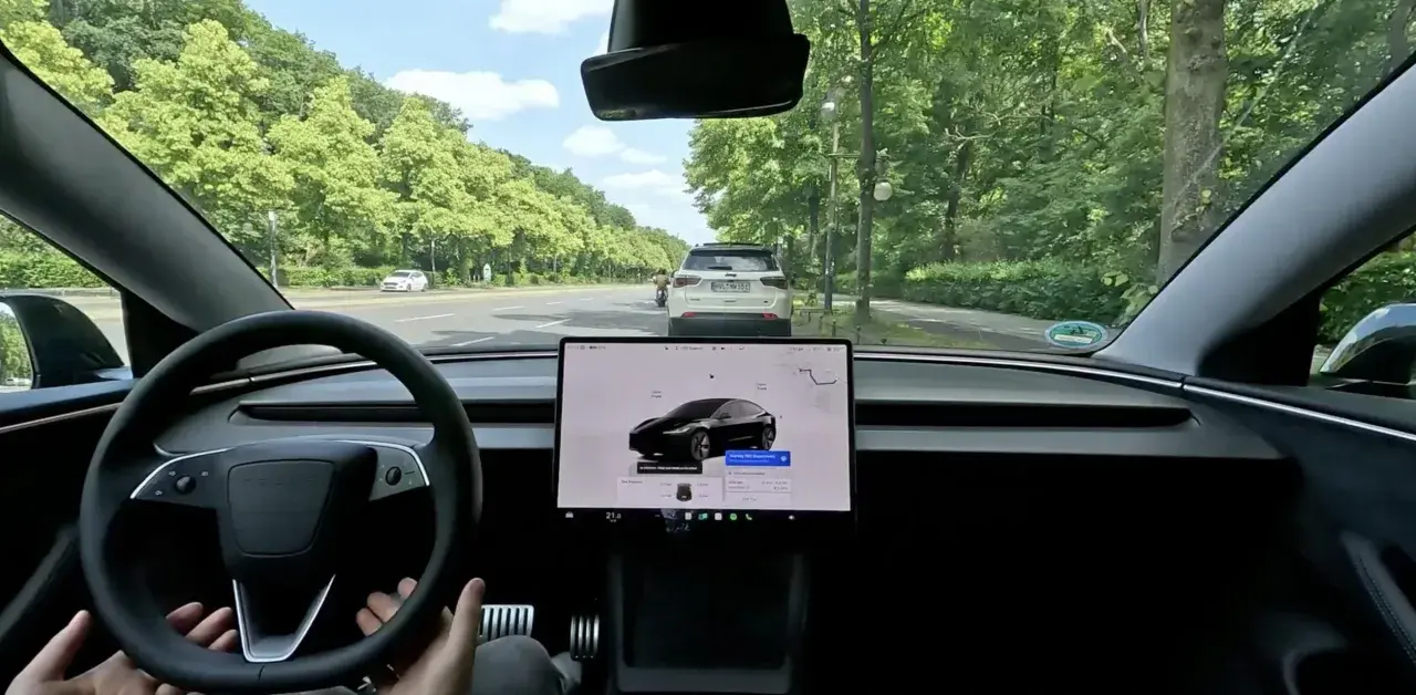 Interno di una Tesla in movimento in Europa, con il sistema FSD attivo. La strada è fiancheggiata da alberi verdi.