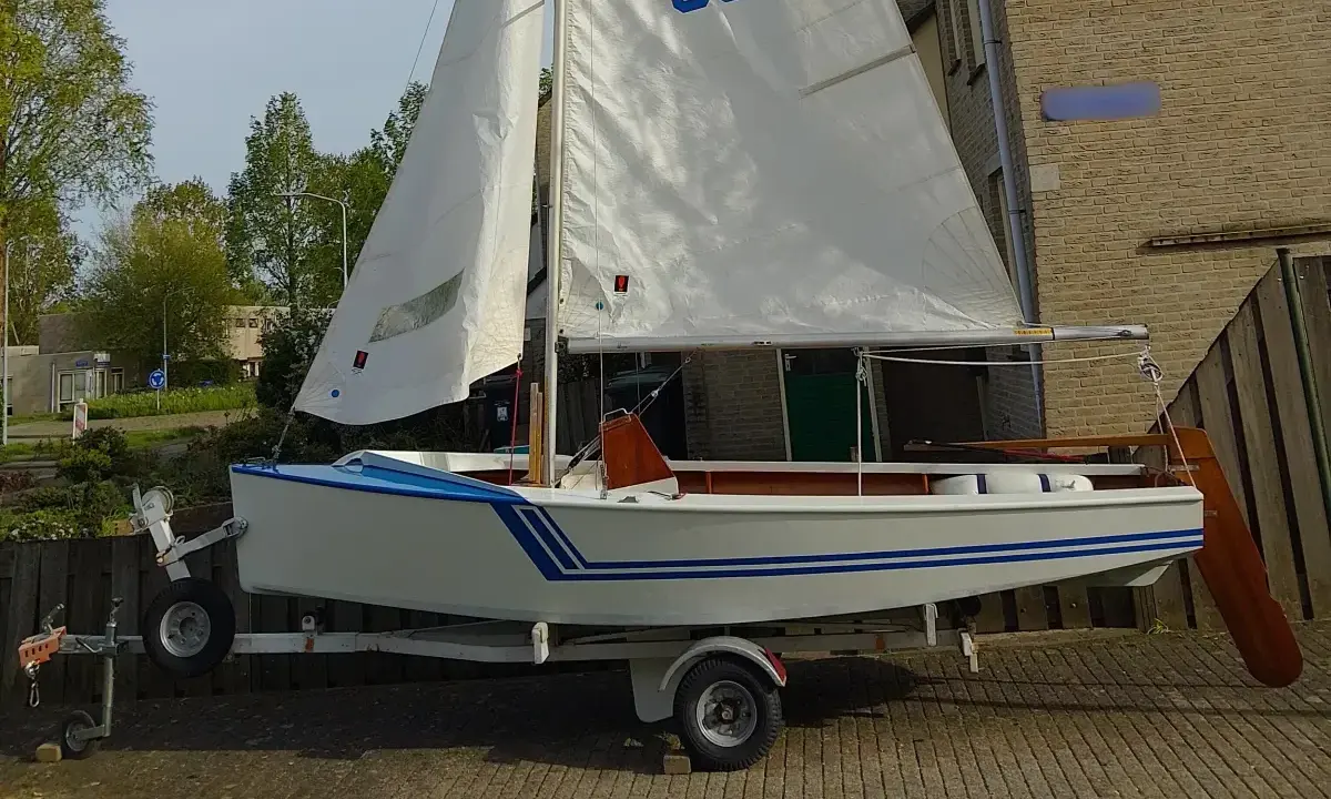 Un petit voilier blanc et bleu, un vaurien bateau prêt pour l'aventure, est sur sa remorque.