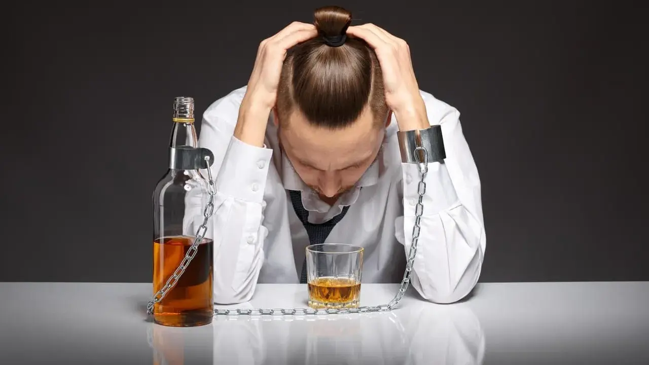 Mężczyzna z rękami na głowie, skuty łańcuchami z butelką i kieliszkiem alkoholu. To symbol walki z alkoholizmem, gdzie pomoc jest kluczowa.