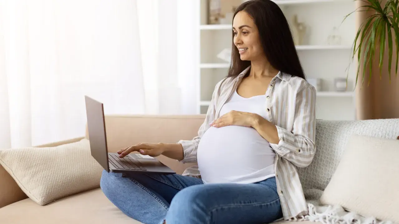 Kobieta w ciąży na kanapie szuka w internecie najdokładniejszy termin porodu, uśmiecha się.