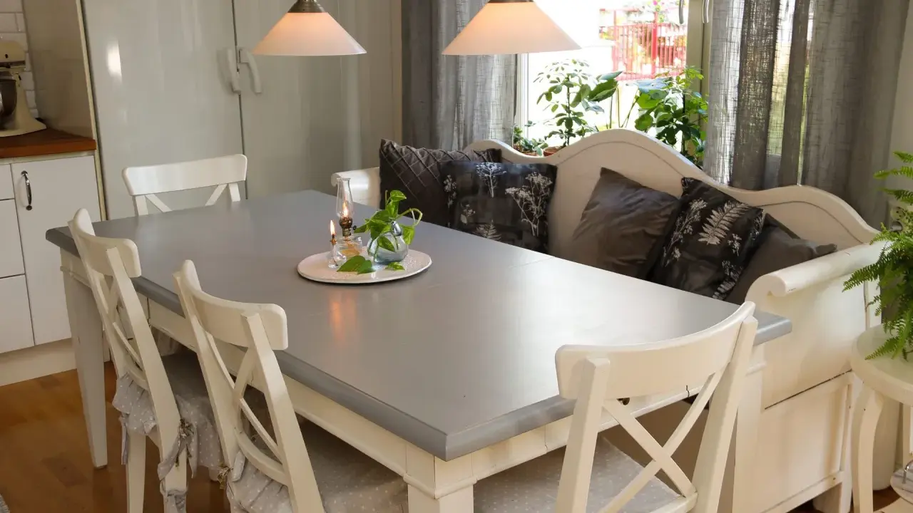 Ett grått bord med vita stolar och en vit bänk med kuddar. Planerar att måla ekbord svart för en modern look.