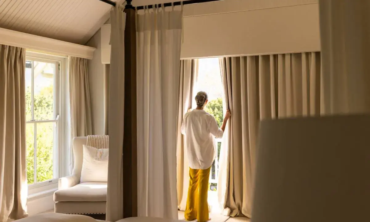 Una mujer abre cortinas para dormitorios, dejando entrar la luz. Se ven camas y un sillón junto a la ventana.