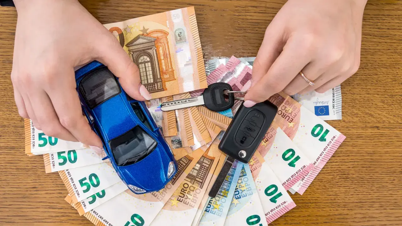 Manos sostienen un coche azul de juguete, llaves y billetes de euro. El coche no arranca, quizás por falta de dinero.