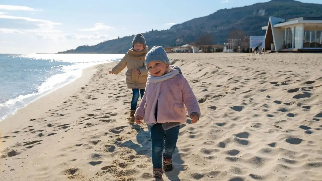 Dwa uśmiechnięte dzieci biegają po piaszczystej plaży. Słońce odbija się od wody, a w tle widać wzgórza i budynek.