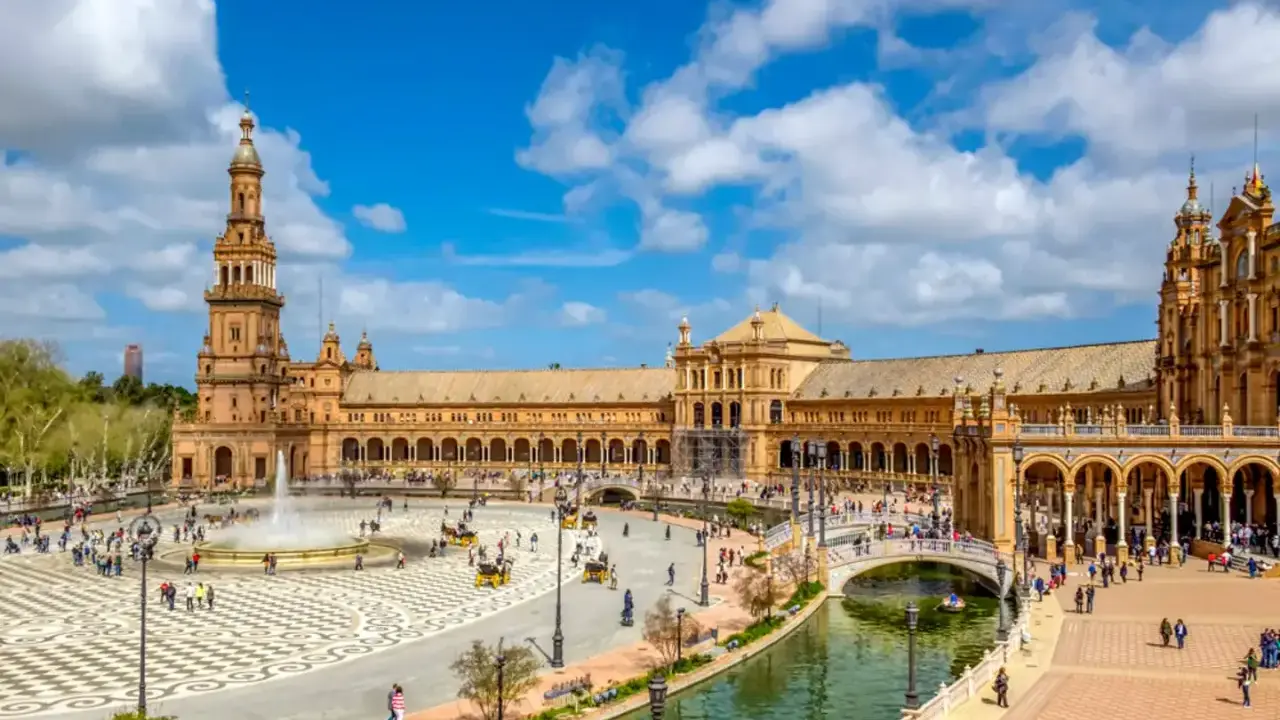Der beeindruckende Sevilla Palast mit seinen Türmen und Arkaden, umgeben von einem Kanal und belebtem Platz mit Brunnen und Kutschen.