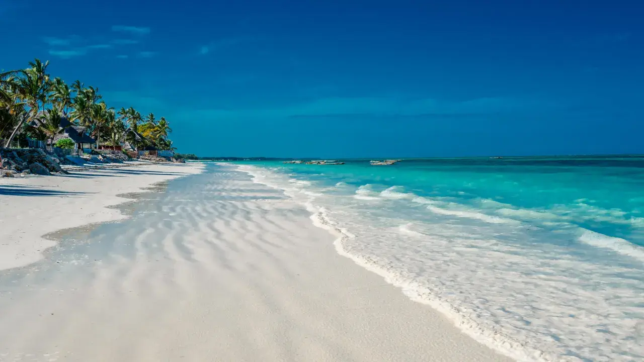 En vit sandstrand med turkost vatten och palmer vid Kendwa, Zanzibar. Båtar guppar i fjärran.