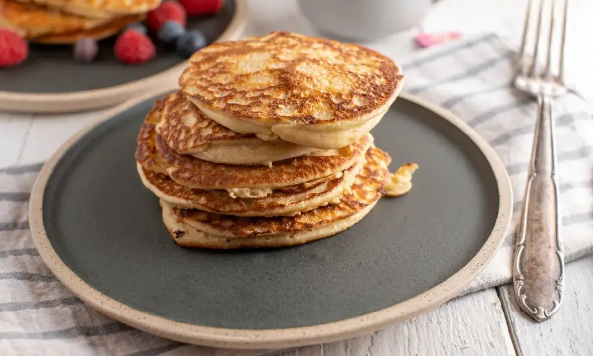 Stos puszystych fit pancakes z odżywki białkowej, ozdobiony malinami i borówkami, czeka na widelcu.