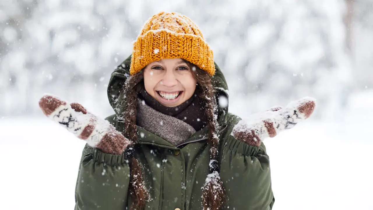 Una chica sonríe felizmente en medio de una nevada, con su gorro naranja y guantes a juego. ¡Bonitas fotos en la nieve!