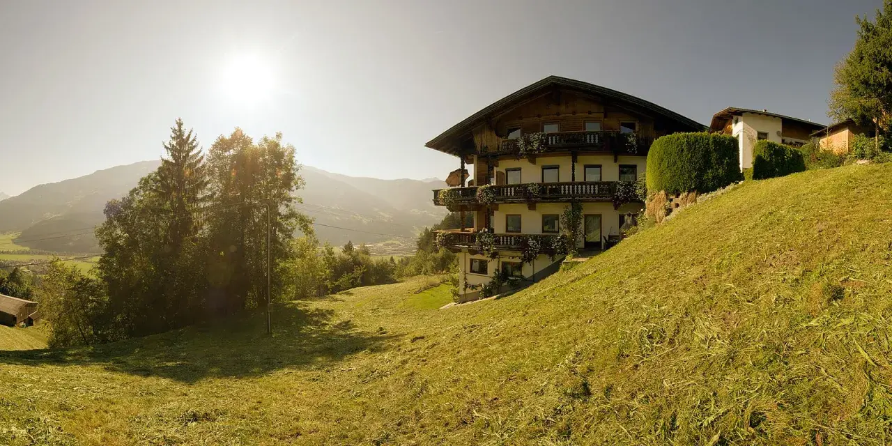 Haus im Alpenvorland mit traditioneller Architektur und Balkonen, umgeben von grünen Wiesen und Bergen.