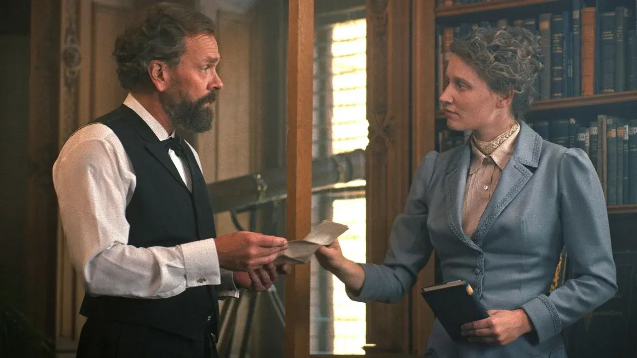 En scen från en alfred nobel film. En man med skägg ger ett brev till en kvinna i en blå dräkt framför en bokhylla.