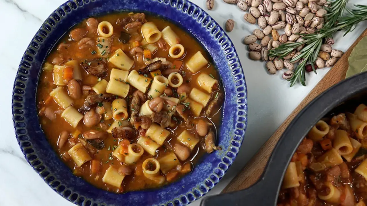Un reconfortante plato de pasta e fagioli en un cuenco azul, con frijoles y romero esparcidos alrededor.