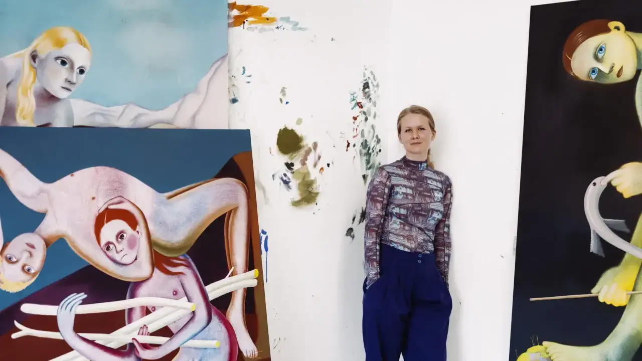 A woman stands in her studio surrounded by her surreal, figurative paintings, showcasing her as one of today's popular contemporary artists.