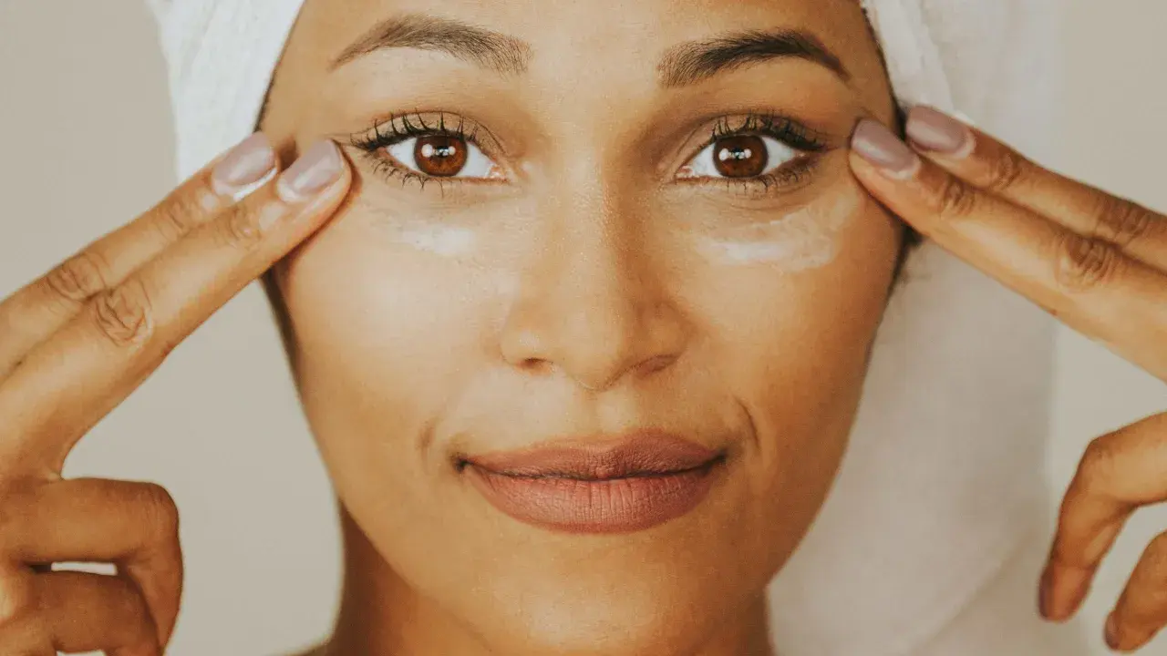 Une femme applique de la crème contour des yeux sur tout le visage, avec une serviette sur la tête.