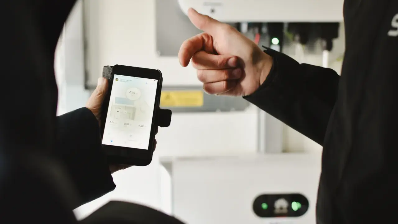 En person håller upp en mobiltelefon med sesol appen som visar solenergi-data. En annan person pekar mot en vit enhet i bakgrunden.
