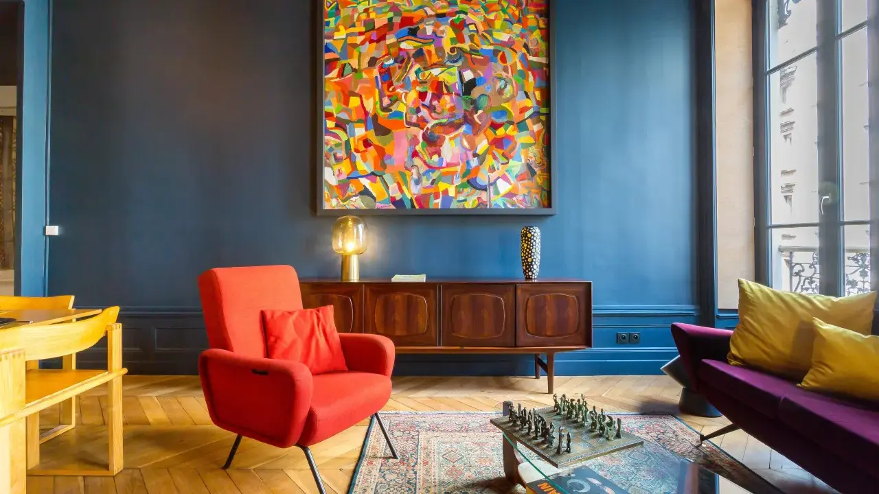 Ein Wohnzimmer mit einem farbenfrohen Gemälde im art déco zeitraum, rotem Sessel, Holzsideboard und einem Schachspiel auf dem Couchtisch.