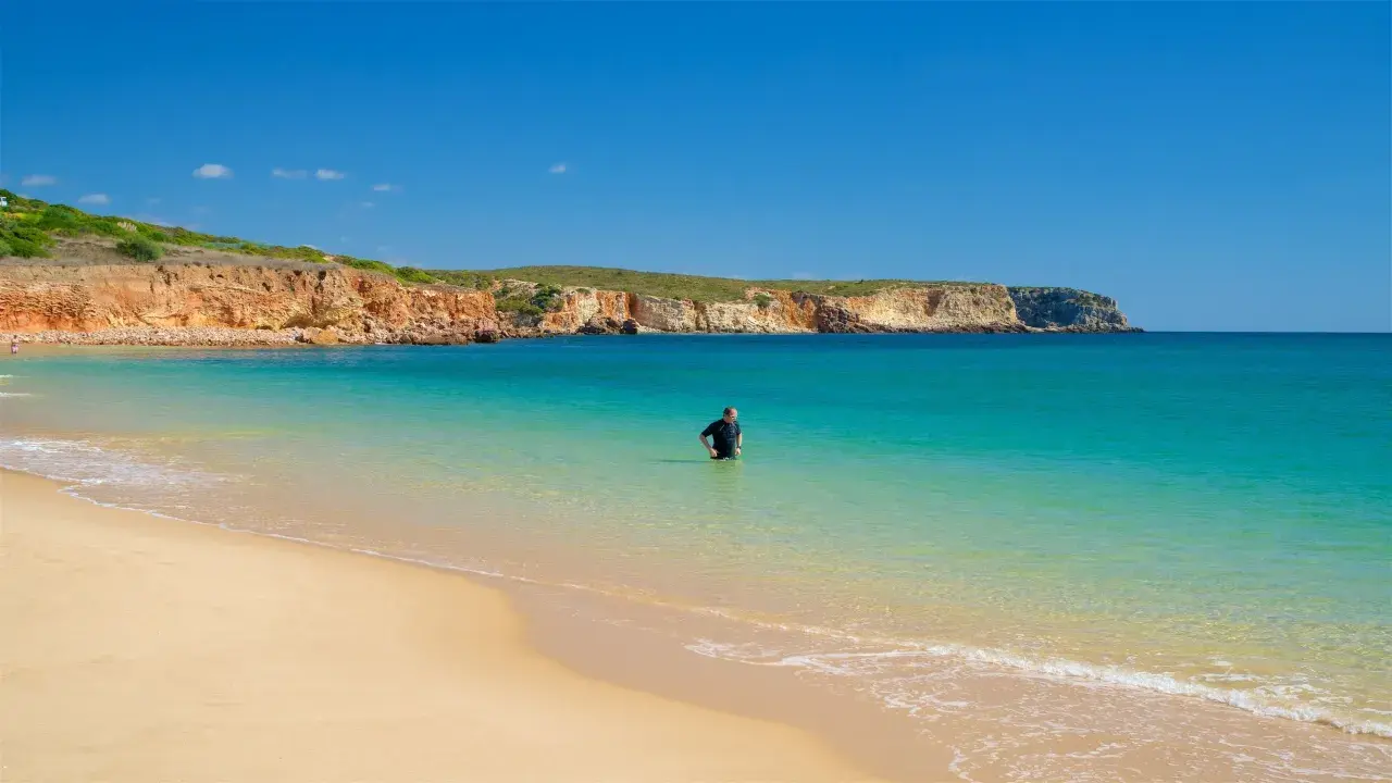 Ein Mann im Neoprenanzug steht im türkisfarbenen Wasser am Praia do Martinhal. Im Hintergrund ragen ockerfarbene Klippen auf.