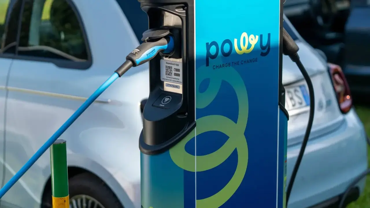 Estación de recarga para coche eléctrico con cable azul conectado. La estación tiene el logo "powy" y el lema "CHARGE THE CHANGE".