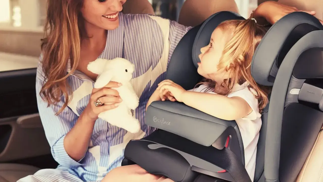 Uśmiechnięta mama z pluszowym zajączkiem przytula córkę w foteliku samochodowym Cybex Pallas 9-36 kg.