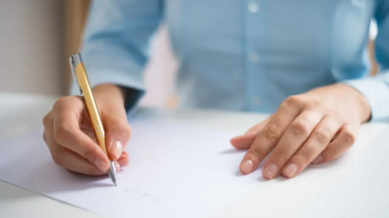 Dłoń pisze złotym długopisem na białej kartce, przygotowując się do egzaminu CAE practice tests.