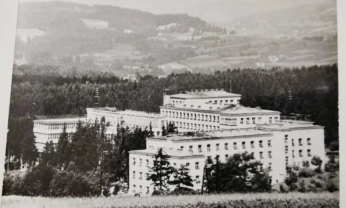 Duży budynek sanatorium w otoczeniu drzew, na tle pagórkowatego krajobrazu.