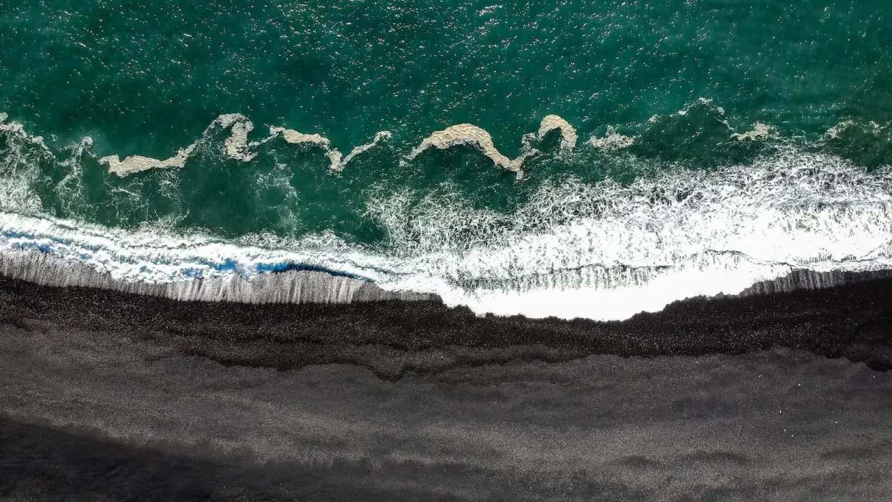 Gran Canaria: Türkisfarbenes Meer trifft auf einen dramatischen schwarzen Strand. Wellen brechen sich auf dem dunklen Sand.