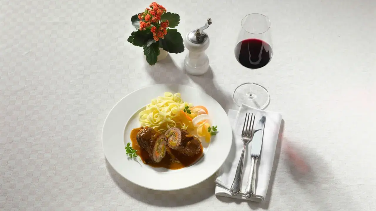 Ein Teller Rouladen mit Bandnudeln und einem Glas feinherb Wein. Dazu ein kleines Blümchen und Besteck.