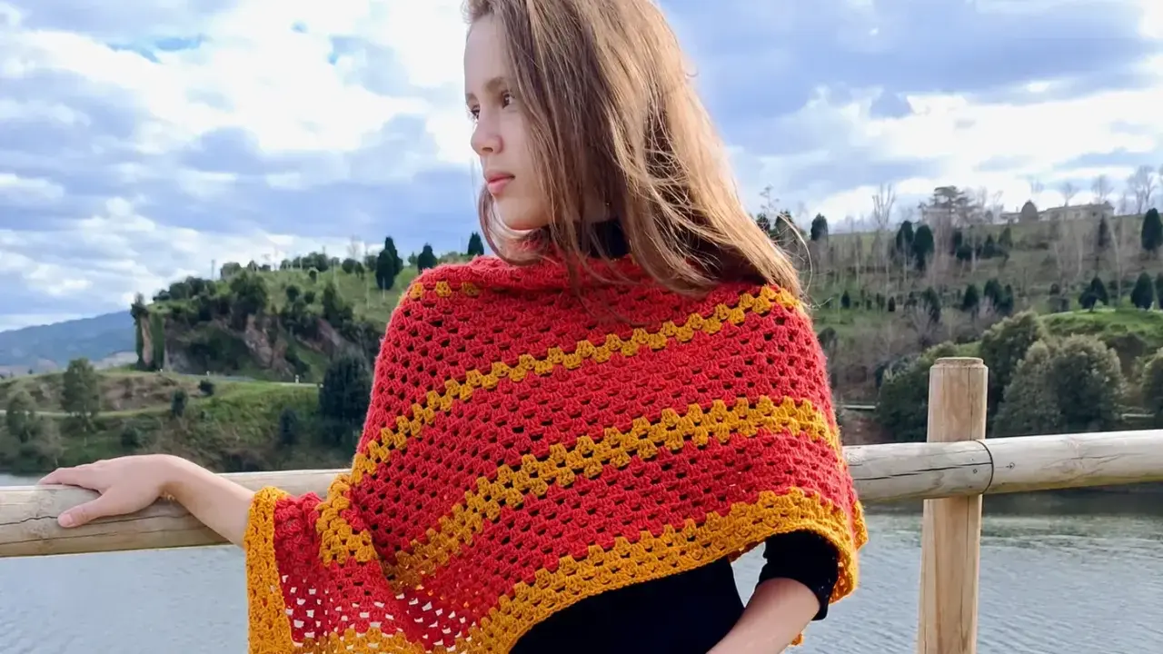 Chica con un pañuelo triangular crochet rojo y amarillo, apoyada en una barandilla de madera junto a un lago.