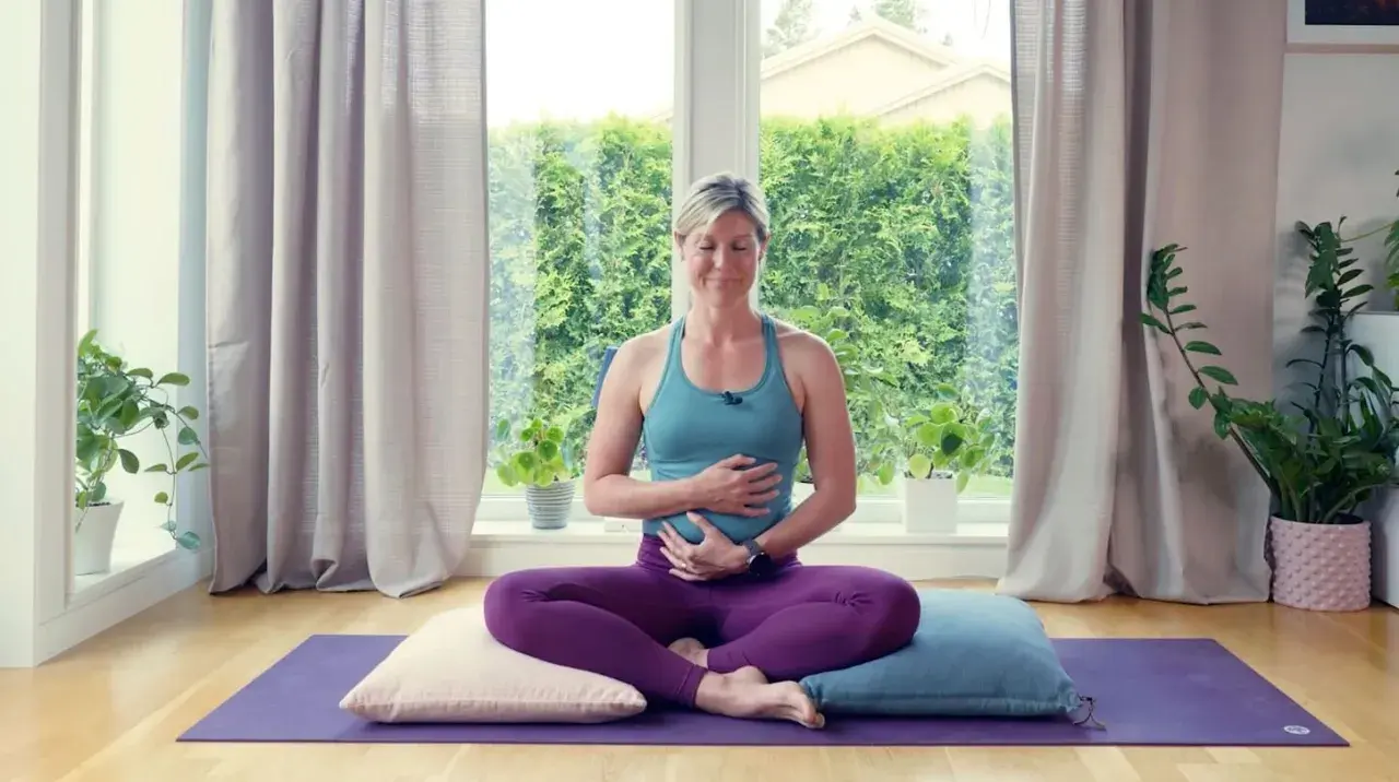 Kvinna i lila kläder sitter i meditationsställning på en yogamatta, med händerna över magen. Hon utövar yoga gravid.