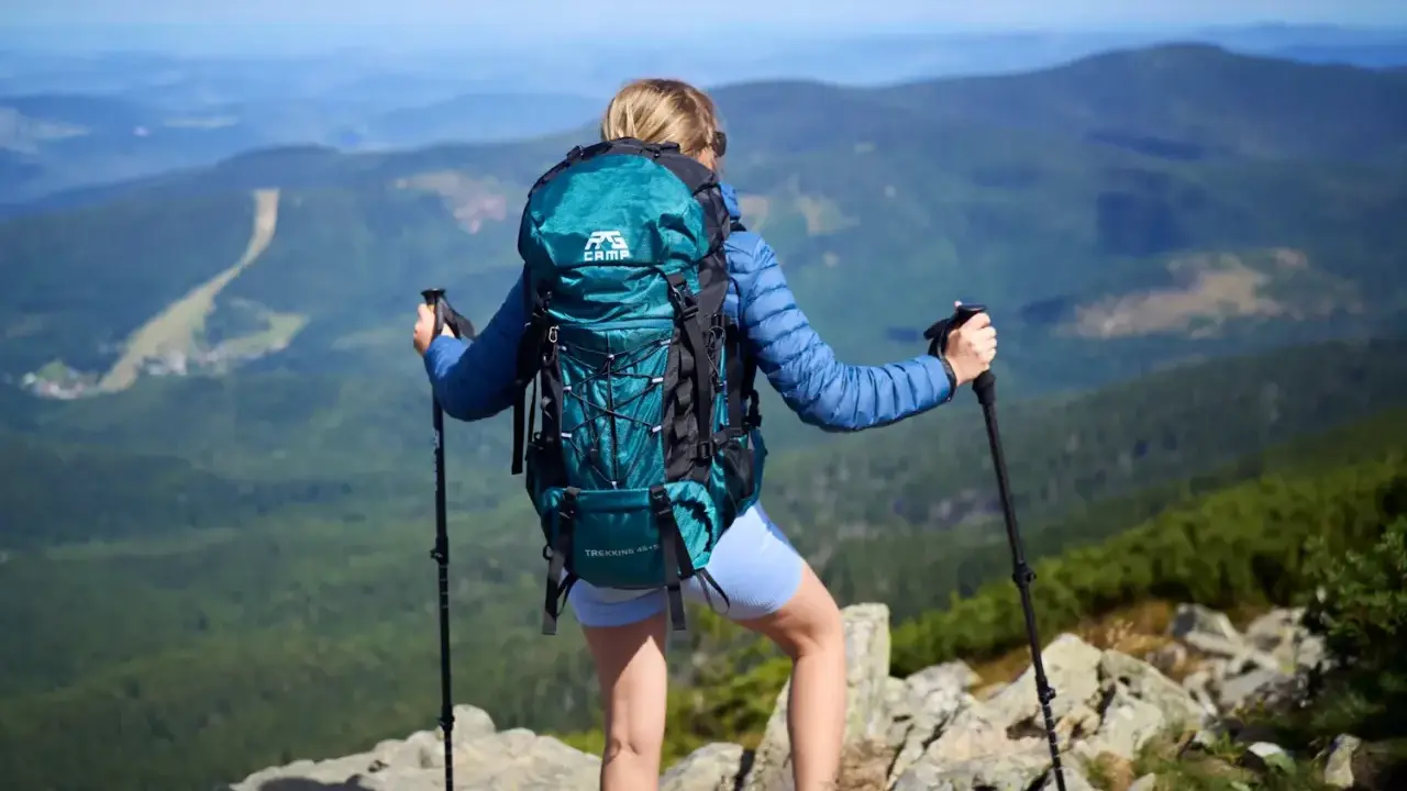 Kobieta z turkusowym plecakiem i kijkami trekkingowymi podziwia widok na góry. Idealny plecak w góry na wyprawę.