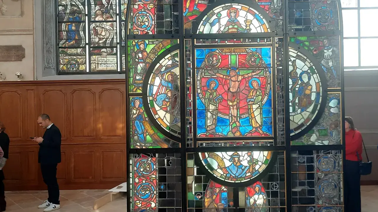 Un vitrail coloré, représentant des scènes bibliques, se dresse dans un intérieur. Un homme consulte son téléphone devant ce chef-d'œuvre.