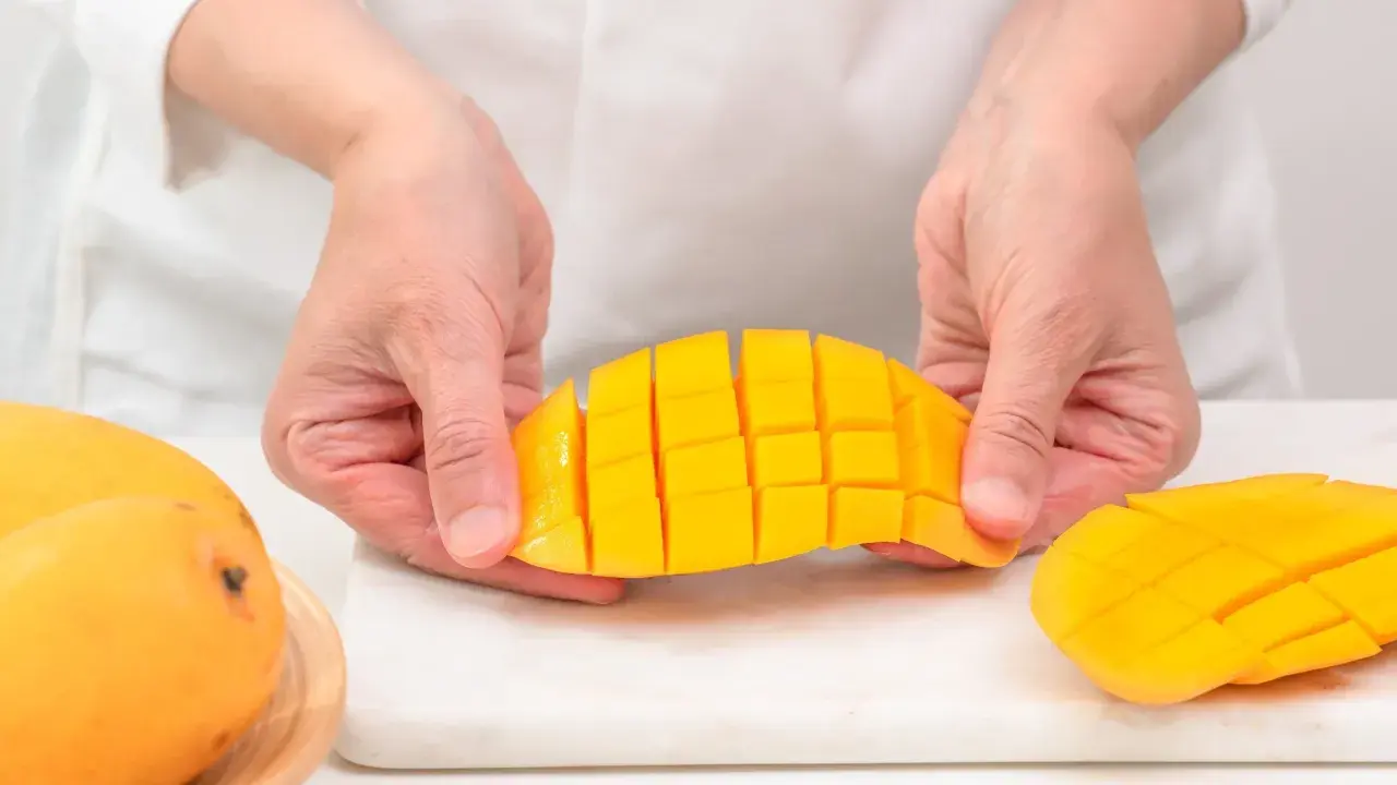Ręce trzymają pokrojone w kostkę mango, pokazując, jak pokroić mango. Obok leżą całe owoce.