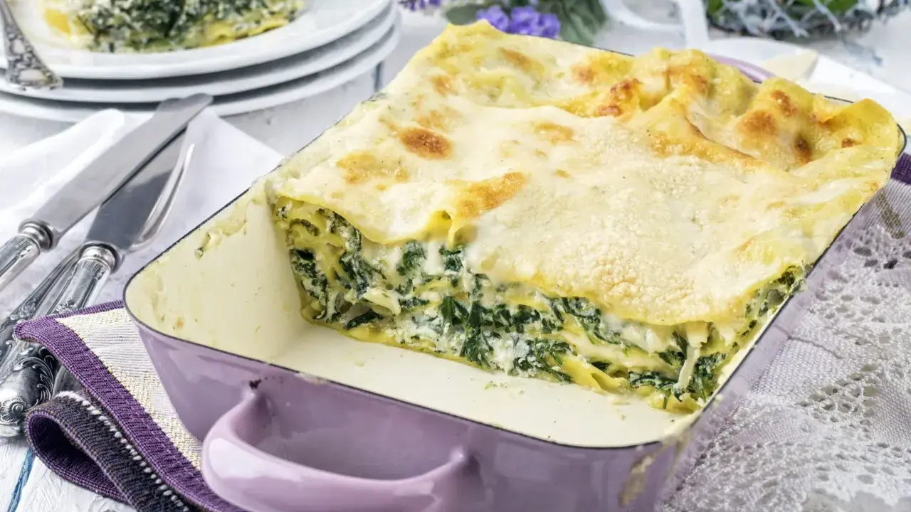 Pyszne lasagne ze szpinakiem w fioletowym naczyniu, gotowe do podania. Obok talerze z porcją i sztućce.