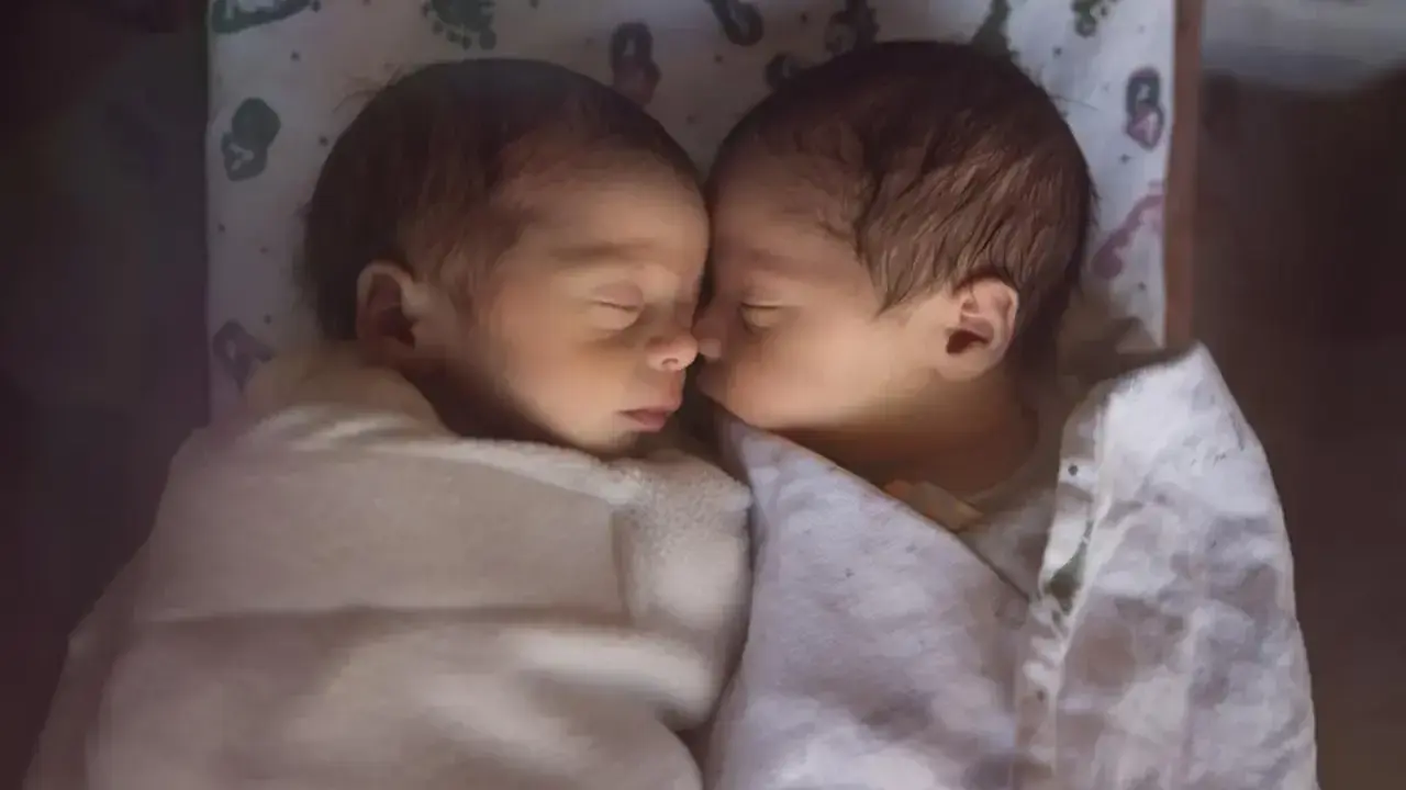 Dwa śpiące niemowlaki, każde w wieku około 2 miesięczne, wtulone w siebie pod białymi kocykami.