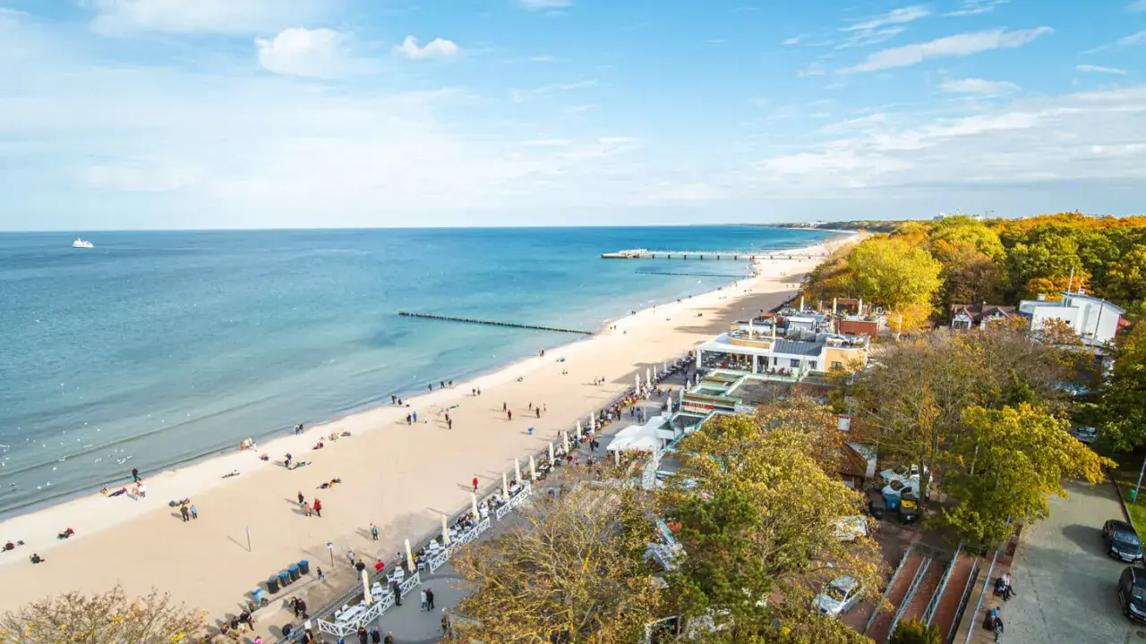 Słoneczna kołobrzeg plaża z ludźmi spacerującymi i odpoczywającymi. W tle morze i molo, a po prawej stronie drzewa w jesiennych barwach.