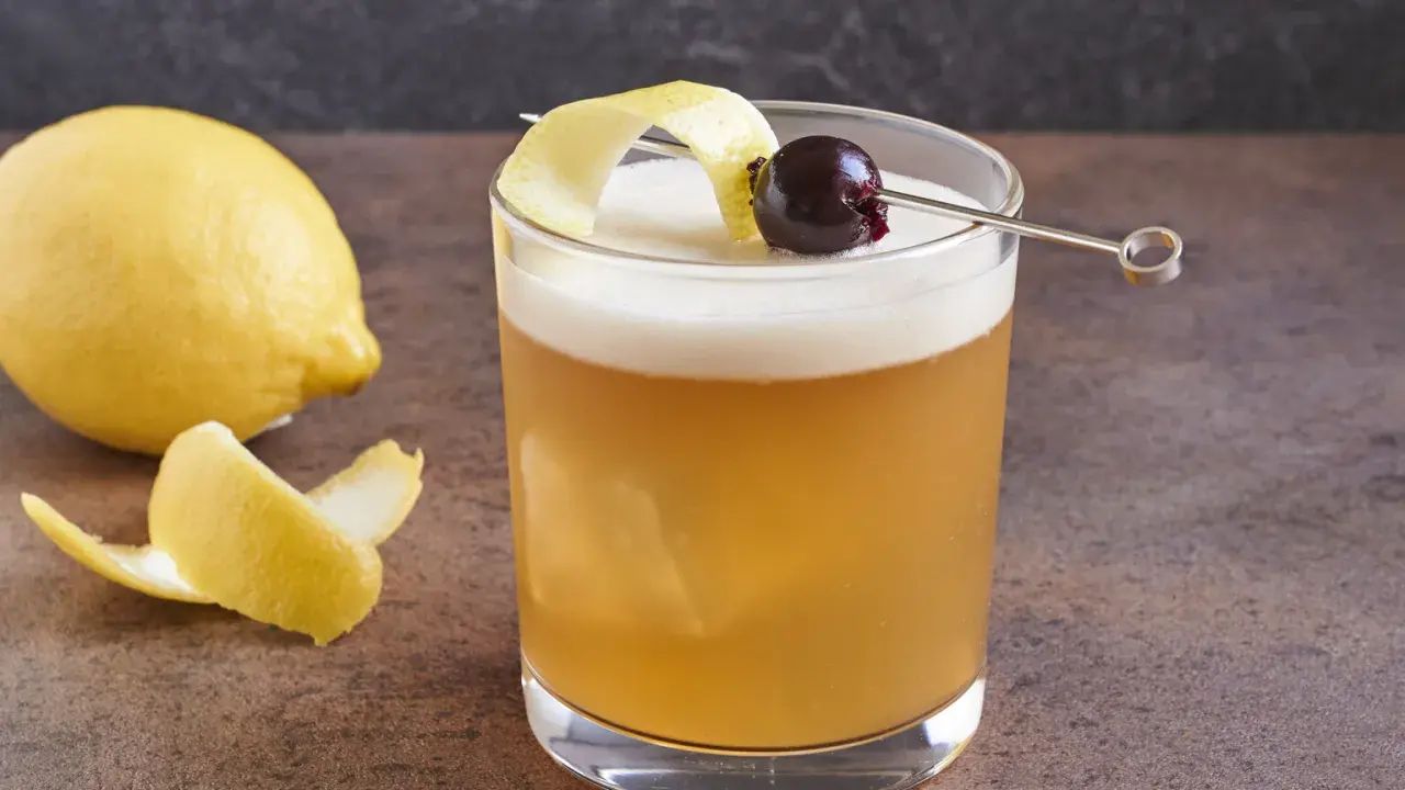 Ein erfrischender Amaretto Sour mit Zitronenzeste und Kirsche, daneben eine Zitrone.