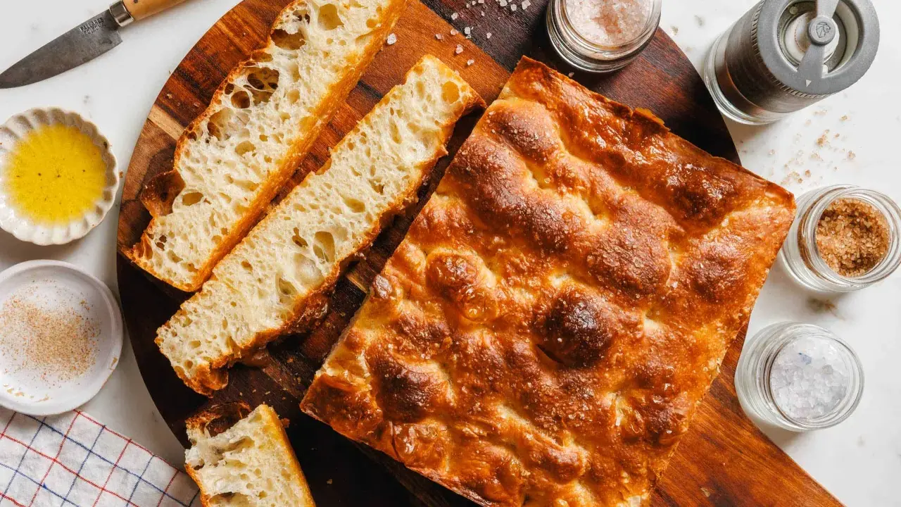 Goldbraune Focaccia mit grobem Salz, bereit zum Belegen. Daneben Olivenöl, Gewürze und ein Messer.