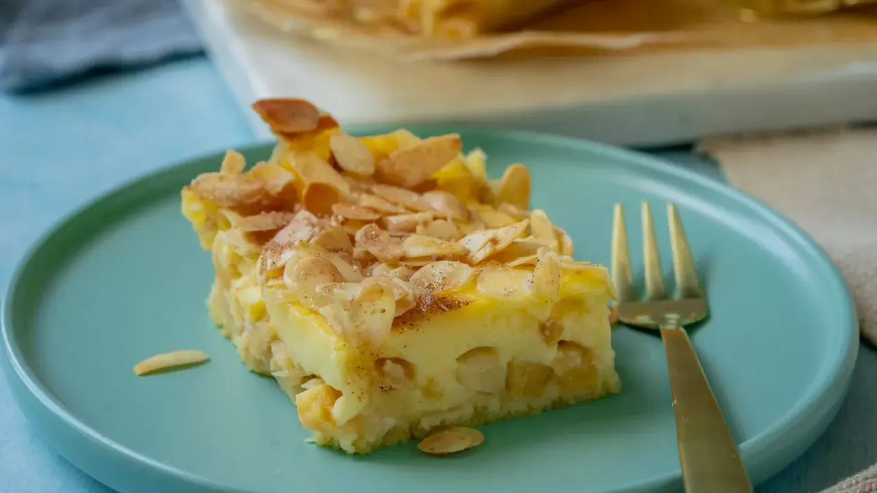 Ein Stück Apfel Schmand Kuchen mit Mandelblättchen bestreut, auf einem türkisfarbenen Teller, daneben eine goldene Gabel.