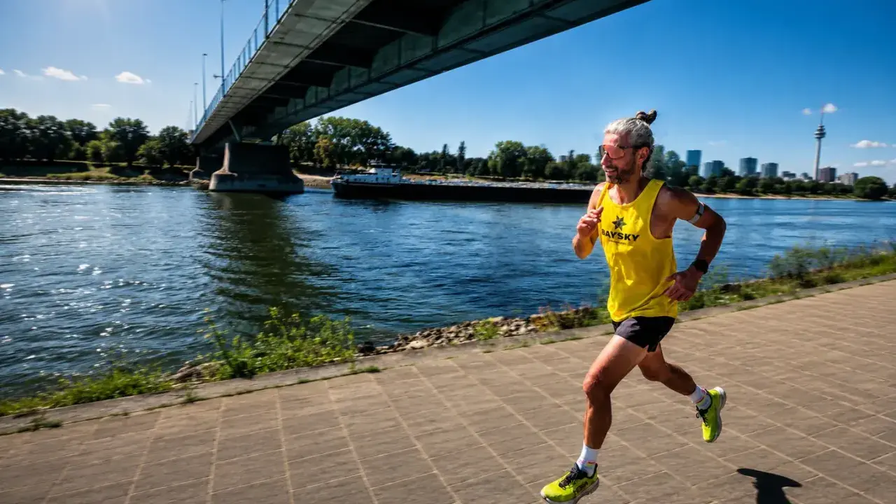 Mann joggt bei 30 Grad am Fluss entlang unter einer Brücke. Sonnenschein, Wasser und eine Skyline im Hintergrund.