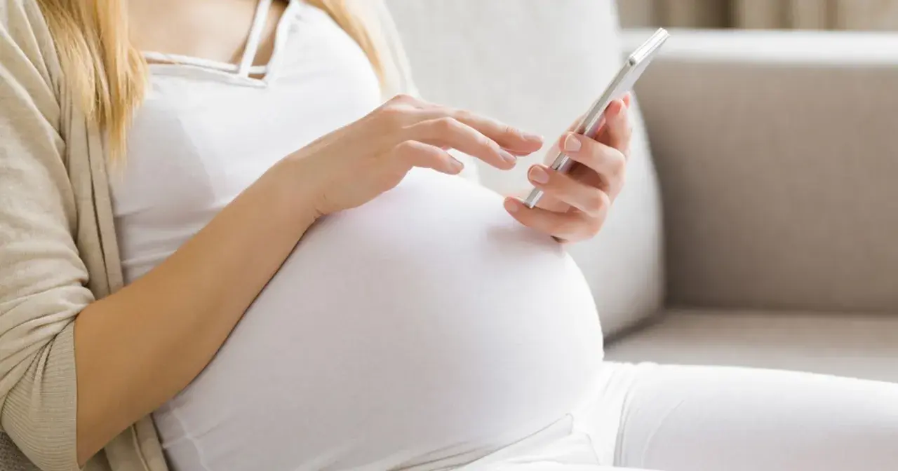 Kobieta w ciąży, w białej bluzce i kardiganie, siedzi na kanapie i korzysta z telefonu. Może sprawdzać zwolnienie lekarskie w ciąży.