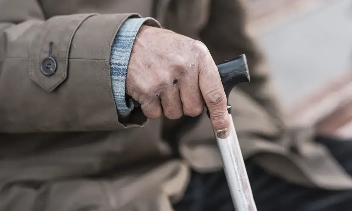 Dłoń starszego mężczyzny z widocznymi oznakami zespołu kruchości, mocno trzymająca laskę.