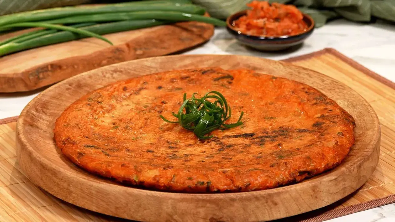 Ein goldbrauner, herzhafter Kimchi-Pfannkuchen, garniert mit Frühlingszwiebeln, liegt auf einem Holzteller. Daneben eine Schale mit Kimchi und grüne Frühlingszwiebeln.