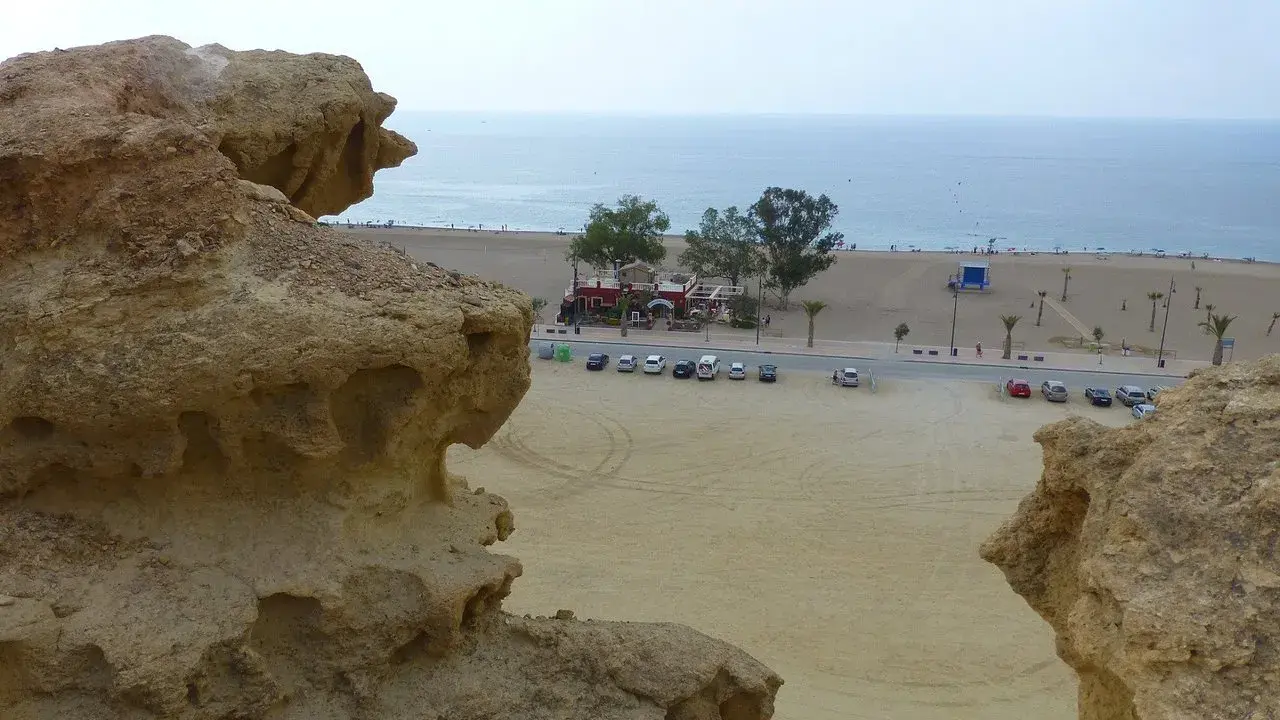 Rocas erosionadas en primer plano, con la playa de Bolnuevo, el mar y un paseo marítimo con palmeras al fondo.