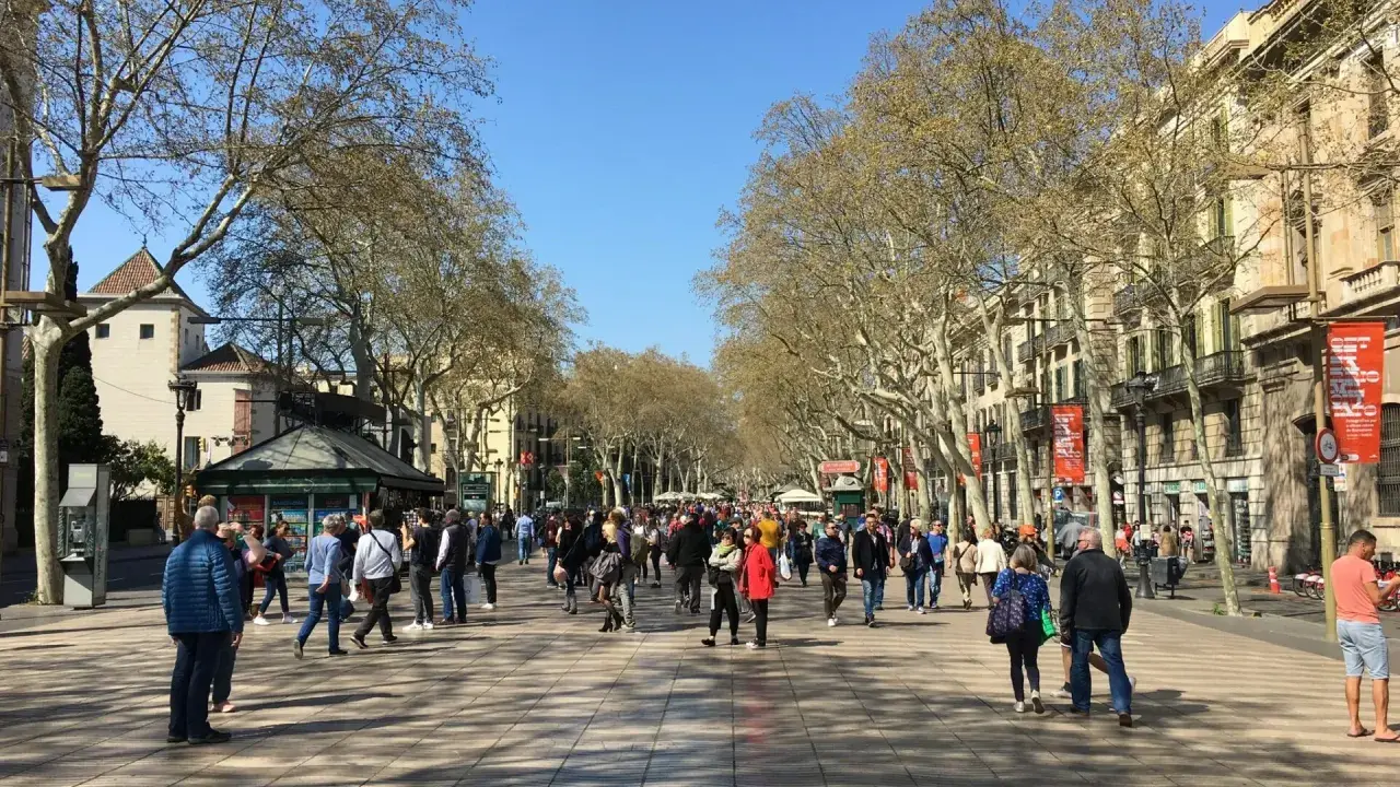 Tłum ludzi spaceruje po słynnej alei La Rambla w Barcelonie, otoczonej drzewami i zabytkowymi budynkami.