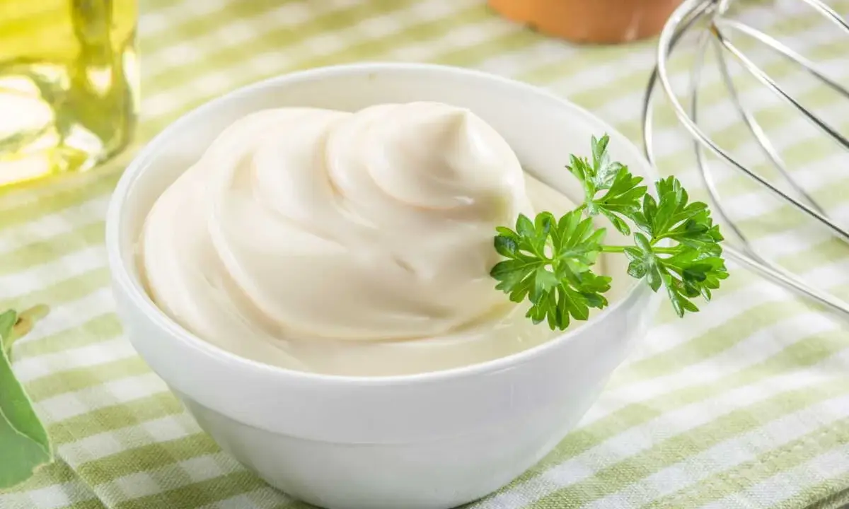 Un bol de mayonnaise maison onctueuse, garni de persil frais. Des œufs et un fouet à proximité suggèrent la préparation.